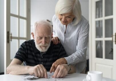 A husband is sitting at a table and enjoying a jigsaw and is being helped by his wife. They are doing the puzzle to help stimulate the mans memory and cognition, to prevent decline and jsut to spend some nice time togeather. Perhaps it is time to think about what care needs this man needs