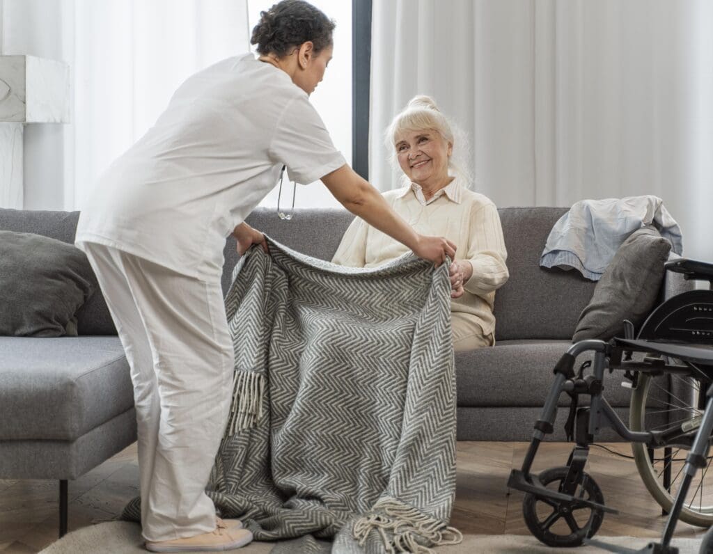 An older adult is being kept warm by a live-in carer offering a blanket onto her legs. The older ladt is happy and comfortable with her carer in the picture and is sitting in her own front room.