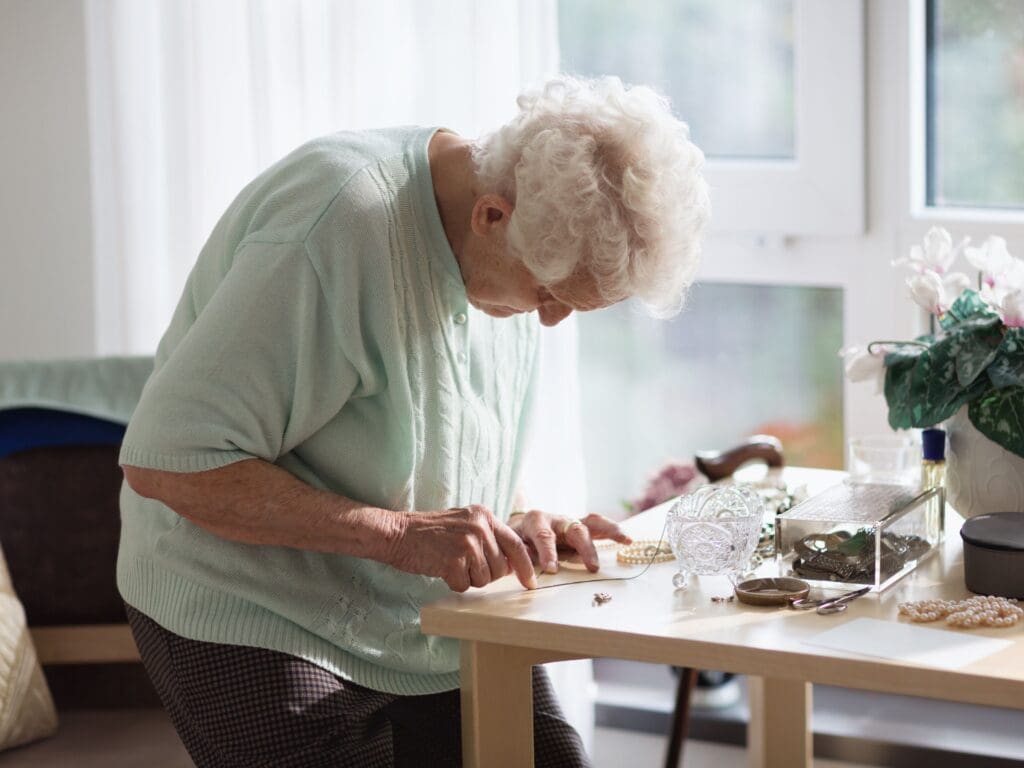 A lady is closly working on what appears to be a haberdashery activity. The lady is staanding next to a table and has lots of natural light in the room to help her with this delicate task