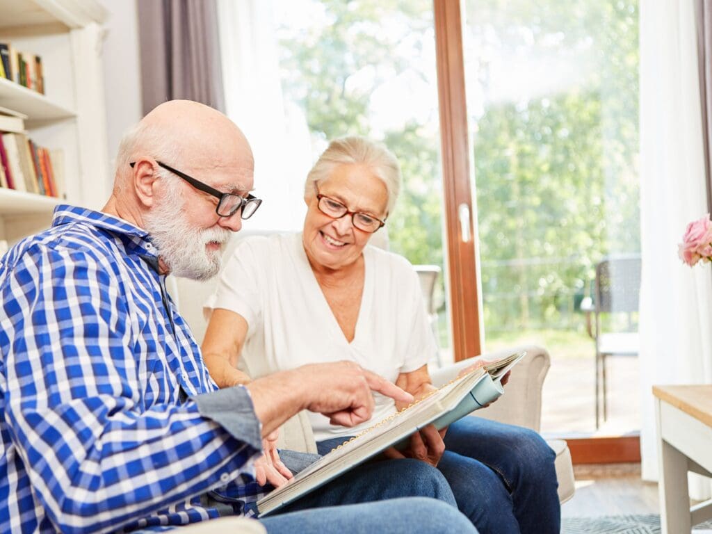 A man with Alzheimers sits next to his wife whilst they both browse a picture album togeather. Looking at pictures will help with his memory and help preserve his family relationships. Some people use electronic photo frames for this too.